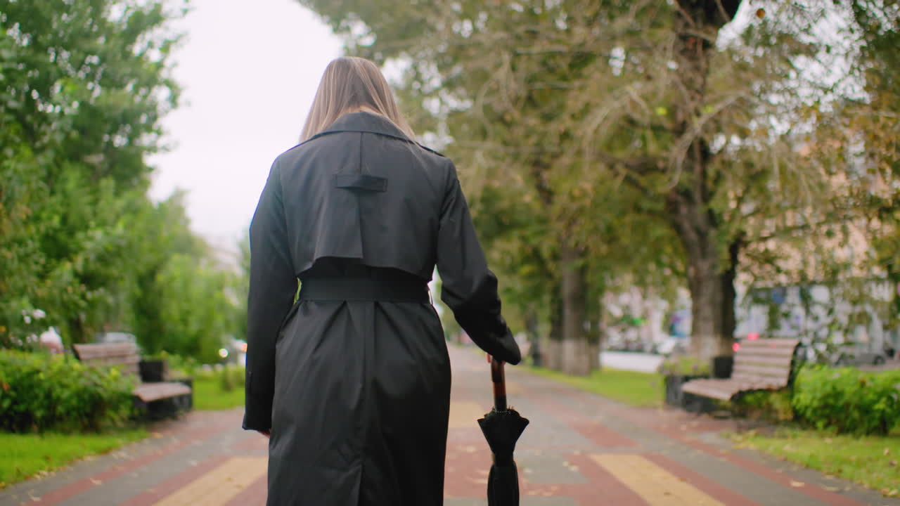 Woman in long black coat walks along tree-lined park path holding closed umbrella on cloudy day, surrounded by benches and greenery, capturing peaceful solitude, urban autumn atmosphere