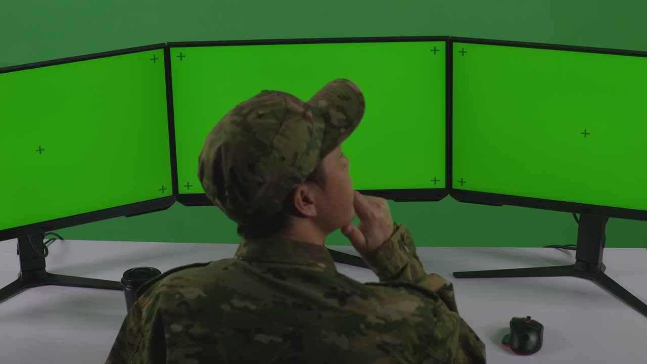 vista trasera de soldado hombre asiático pensando en algo mientras trabajaba con simulacro de múltiples monitores de computadora en el estudio de pantalla verde