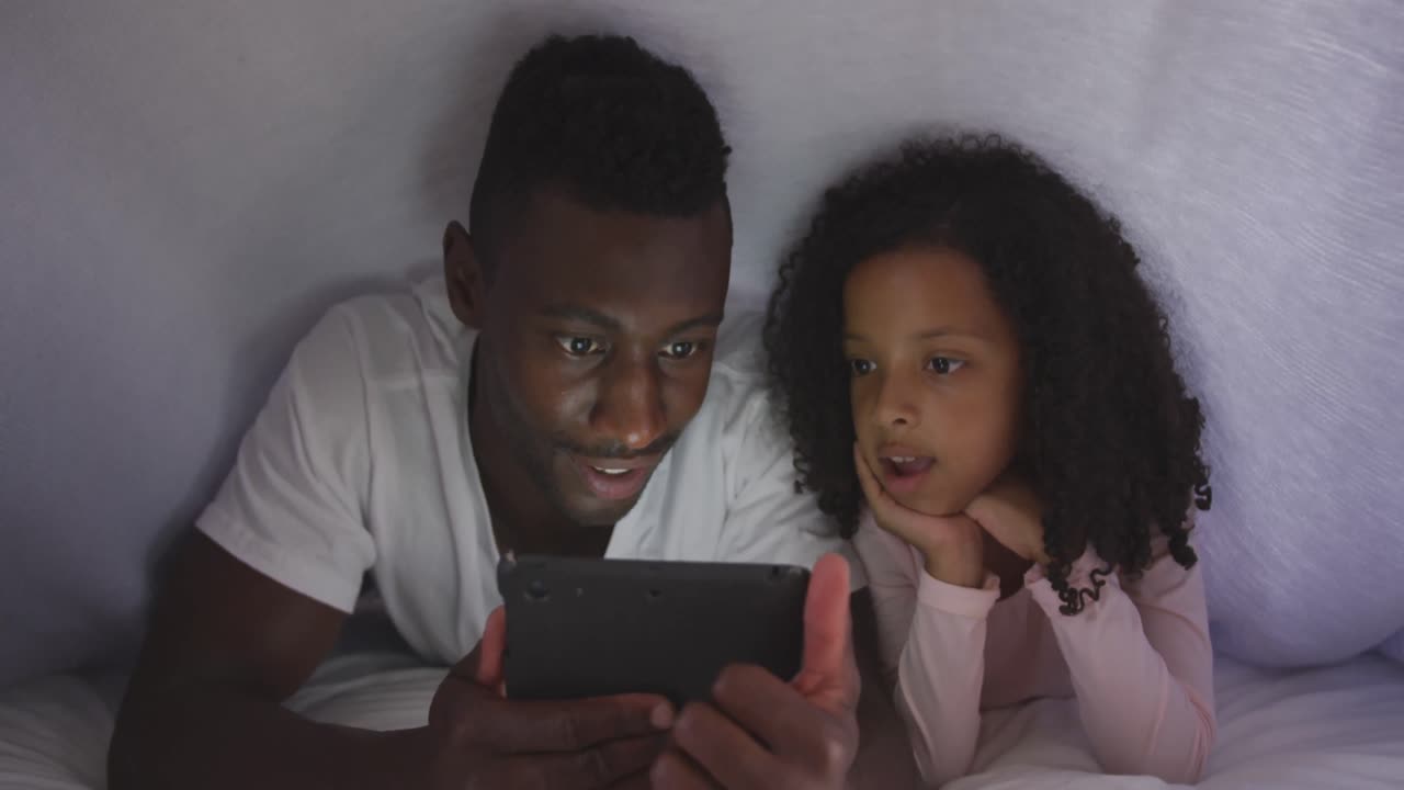 African american father and daughter looking at phone in bed