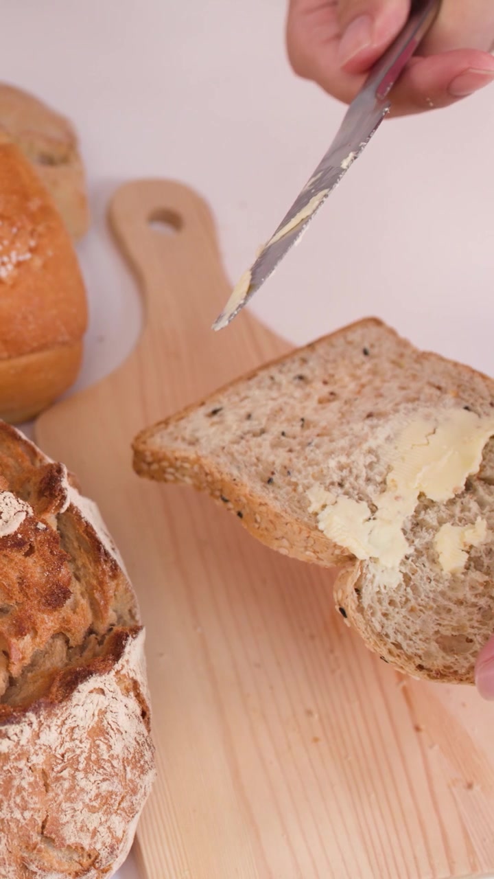 Close-up of hand spreading butter on multigrain slice amidst sourdough, baguettes, and croissants.