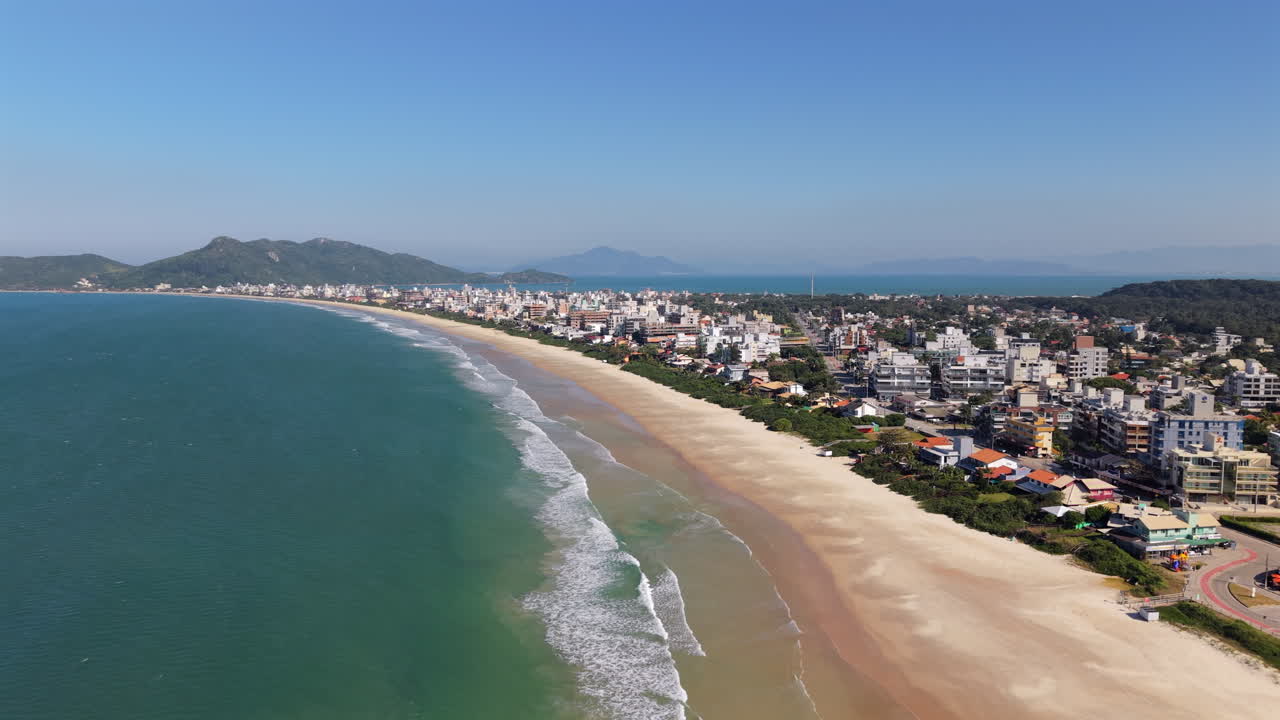 Wide sandy beach of Mariscal, Santa Catarina, Brazil, showing rolling waves, a long coastline, nearby urban buildings, lush greenery, and distant mountains under a clear blue sky, drone pull out