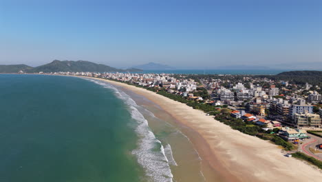 wide sandy beach of mariscal, santa catarina, brazil, showing rolling waves, a long coastline, nearby urban buildings, lush greenery, and distant mountains under a clear blue sky, drone pull out