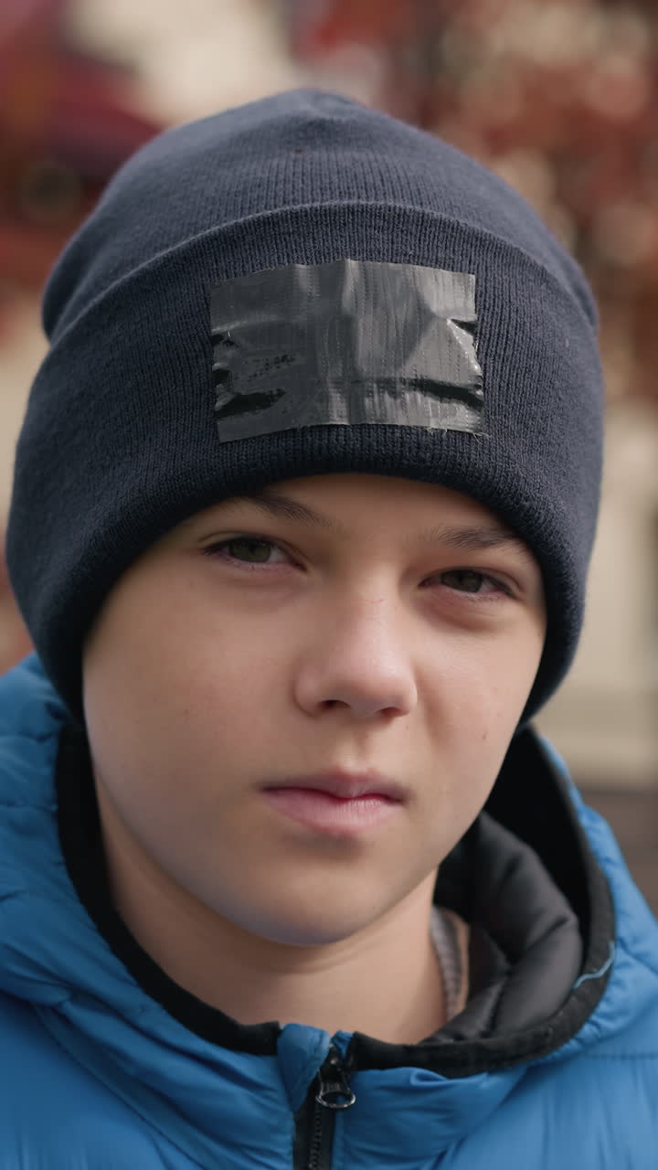 primer plano de un niño con gorra negra y chaqueta azul al aire libre, mirando tranquilo y reflexivo, con una casa borrosa de techo rojo y árboles de otoño vibrantes en el fondo