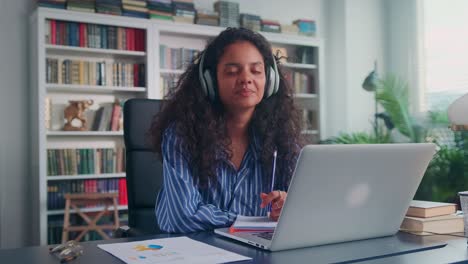 young indian woman student in wireless headphones sits at desk in home office
