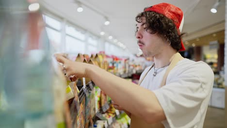 side view of a brunette guy with curly hair chews a bun and rearranges goods on the shelves during his work in a supermarket