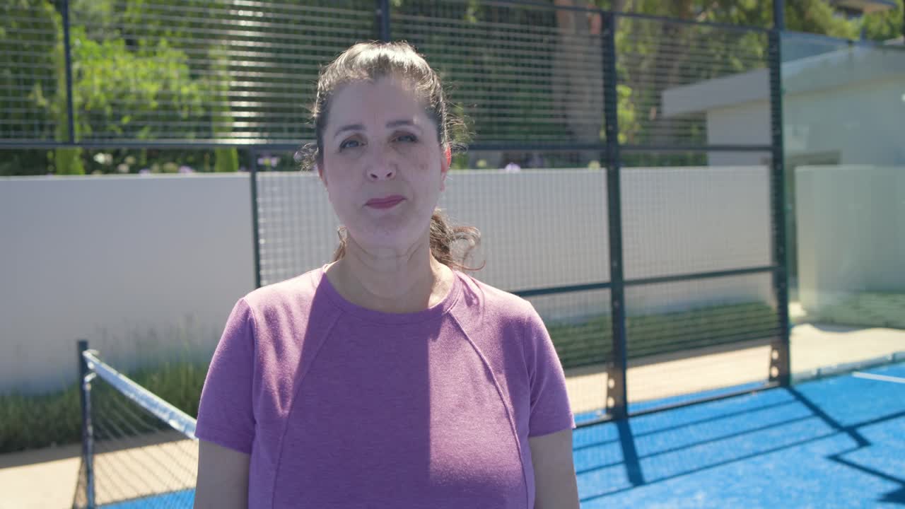 Wanita senior Afrika-Amerika berbaju ungu meletakkan raket pickleball di bahu di lapangan biru sambil tersenyum.