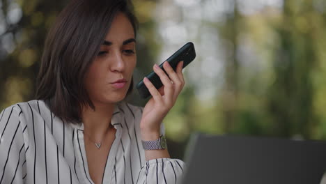 Multitasking-hispanic-businesswoman-using-voice-recognition-function-on-a-smartphone-standing-near-desk-and-typing-on-the-laptop.-Arabian-female-entrepreneur-sending-a-vocal-message