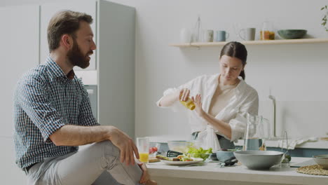 happy woman standing seasoning salad, while man sitting on stool telling her something funny and make her laugh