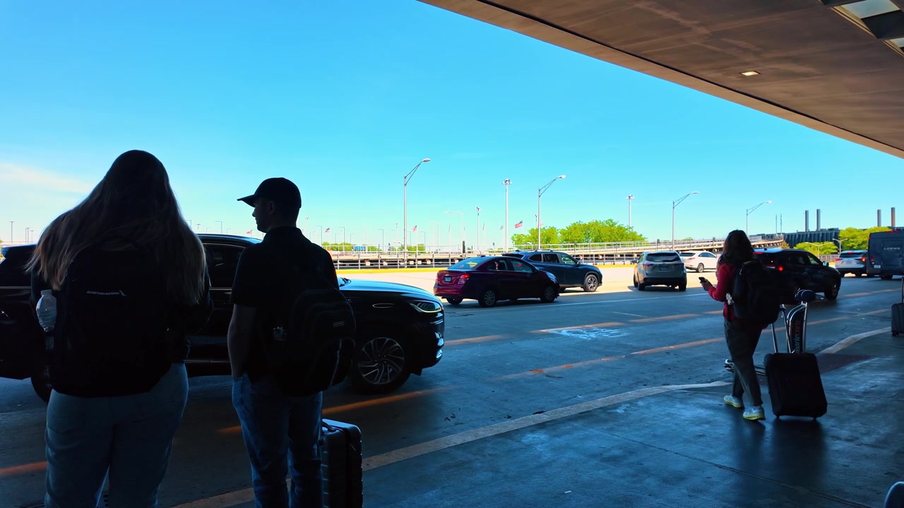Chicago, USA, 27. maj 2026 : Passagerer venter ved lufthavnens afhentningszone ved kantstenen. Folk står med bagage under et terminaloverdækning, mens biler kører forbi på lufthavnsvejen.