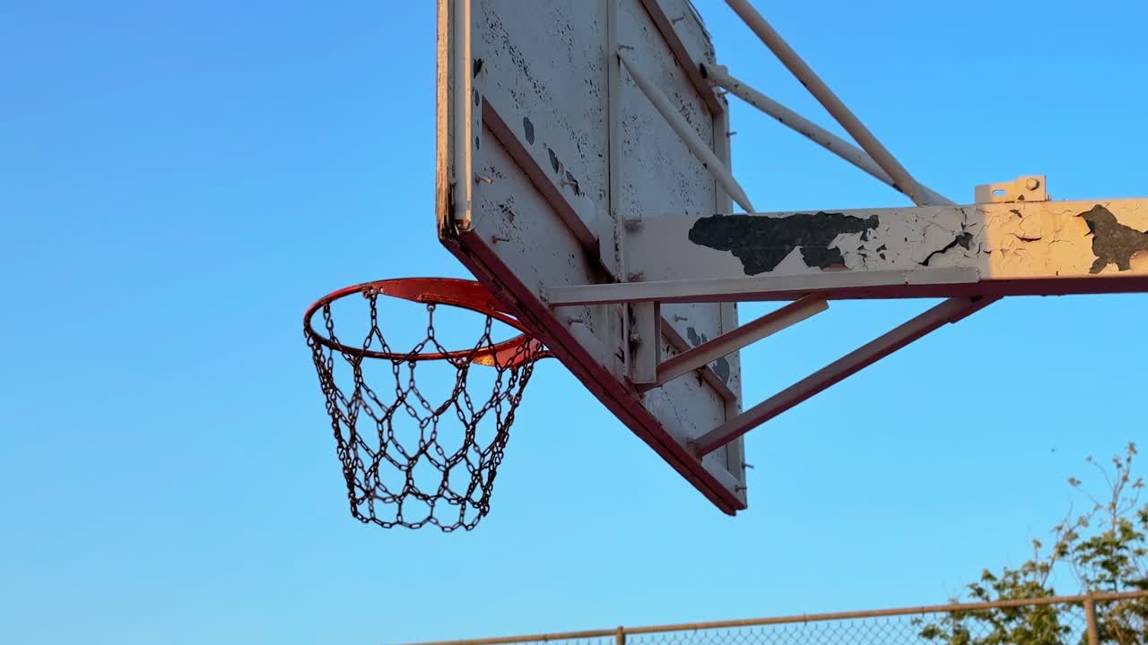 Detailed view of a rusty chain net basketball hoop under a blue sky. Ideal for urban sports, motivation, street culture, and cinematic B-roll footage.