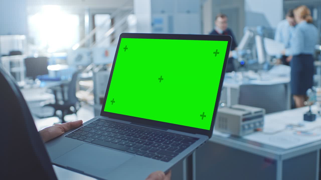 el ingeniero sostiene una computadora portátil de pantalla de maqueta verde. en el fondo, un equipo de ingenieros industriales trabaja con un brazo robótico funcional en una moderna instalación de investigación brillante