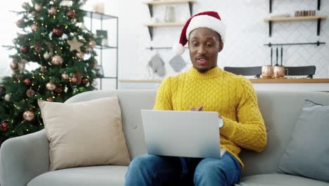 Fröhlicher-Gutaussehender-Kerl-Mit-Weihnachtsmütze-Videochattet-Am-Computer,-Während-Er-Zu-Hause-In-Der-Nähe-Des-Geschmückten-Glühenden-Neujahrsbaums-Sitzt