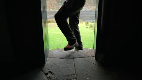 A-man-with-dreadlocks-and-wearing-hiking-gear-sits-on-a-step-in-a-narrow-doorway-of-a-bothy-in-the-Highlands-of-Scotland-whilst-enjoying-the-view-outdoors