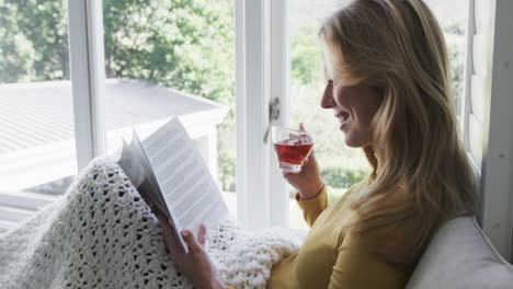 Happy-biracial-woman-sitting-next-to-window-drinking-tea-and-reading-book-at-home,-slow-motion