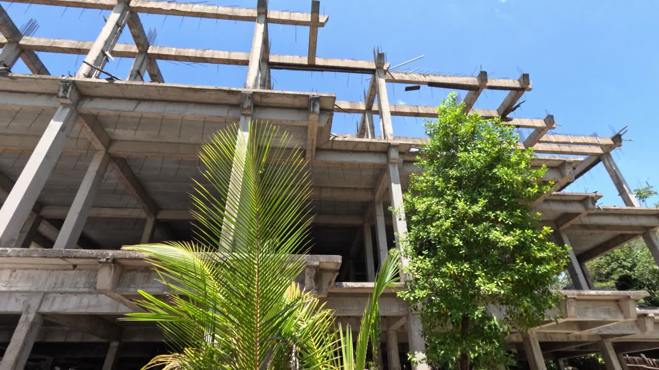 Unfinished concrete hotel structure surrounded by palm trees, bright daylight, upward camera pan movement