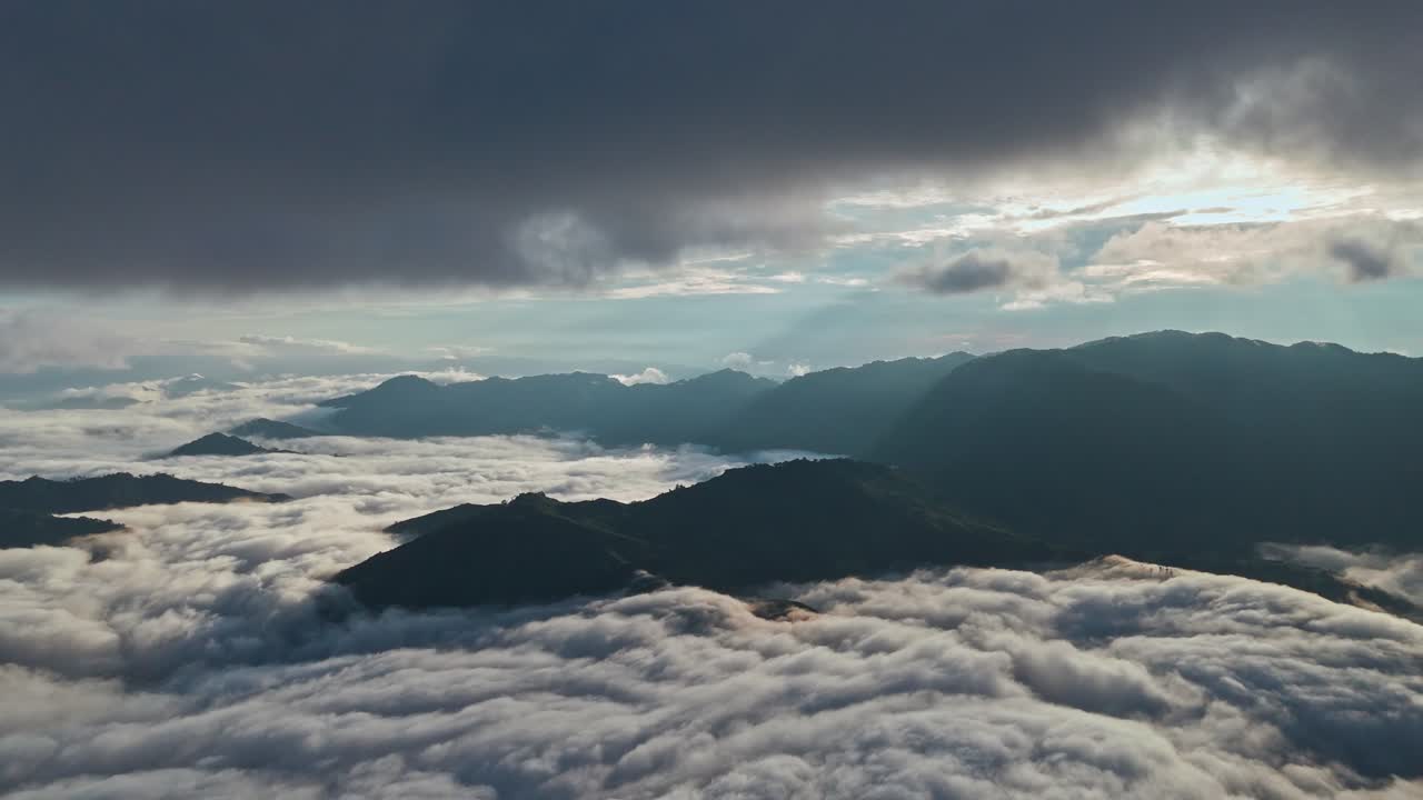 Wide aerial view of mountains rising above a sea of clouds, with sunbeams breaking through dark storm clouds in early daylight, creating a cinematic, atmospheric landscape scene