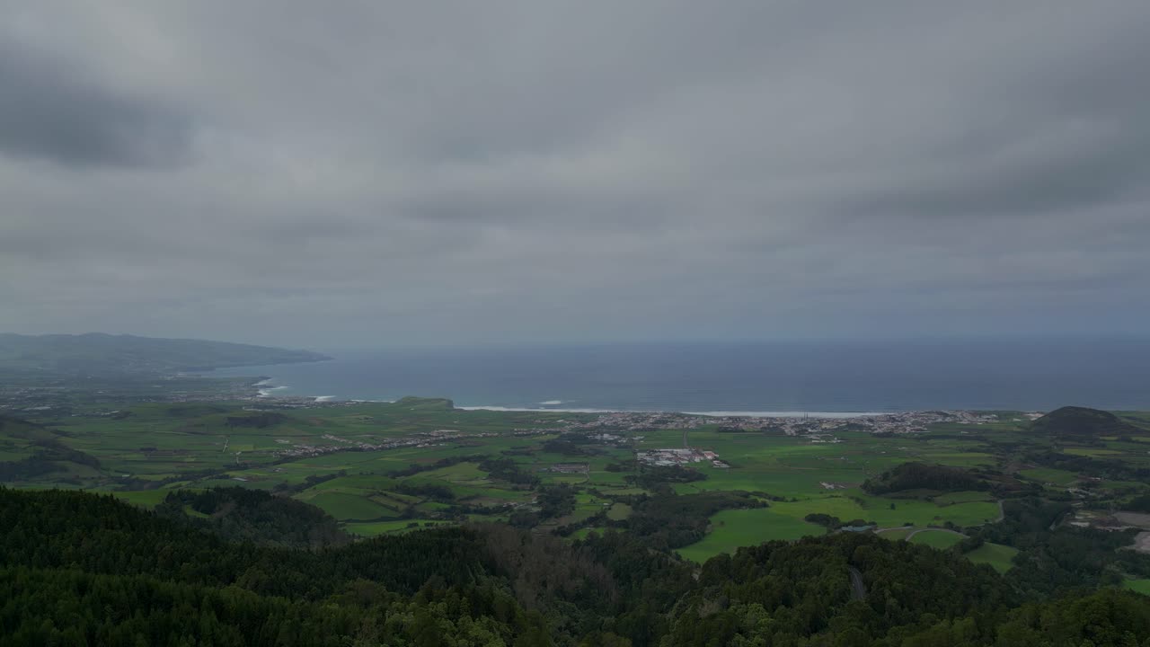 Expansive view of a cloudy coastal landscape in the Azores, Portugal. Rolling green fields meet the blue ocean under a dramatic overcast sky, blending natural beauty with atmospheric tranquility.