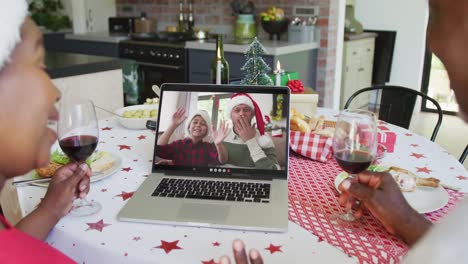 African-american-couple-with-wine-using-laptop-for-christmas-video-call-with-happy-family-on-screen