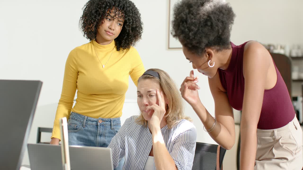 Colegas de trabalho femininas diversas em uma mesa, uma delas de blusa bordô apontando uma caneta para o laptop enquanto sentada revisando conteúdo.