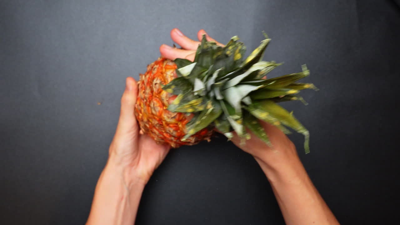 Person twisting a pineapple to prepare it for cutting in a dark setting. Two hands are holding a ripe pineapple and twisting it to prepare for slicing on a black surface