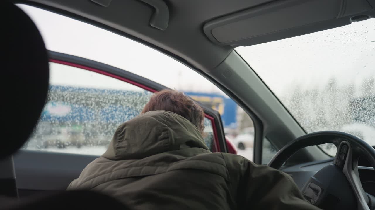Back view of man in winter jacket leaving car interior and shutting door as snow falls outside, wet window shows raindrops with blurry cityscape in background