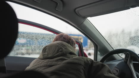 back view of man in winter jacket leaving car interior and shutting door as snow falls outside, wet window shows raindrops with blurry cityscape in background