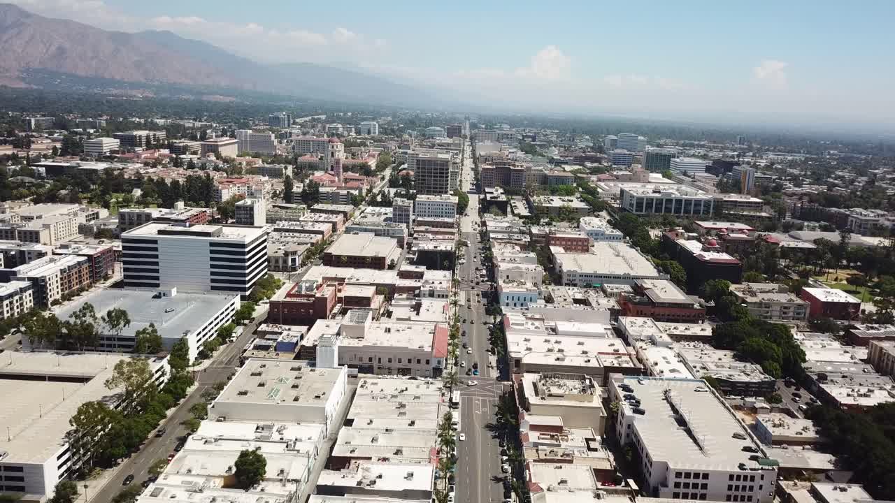 Pasadena, California. Auto che percorrono un lungo viale cittadino fiancheggiato da edifici bassi, uffici e alberi. I monti San Gabriel sono visibili in lontananza sotto un cielo sereno. Ampia ripresa aerea all'indietro