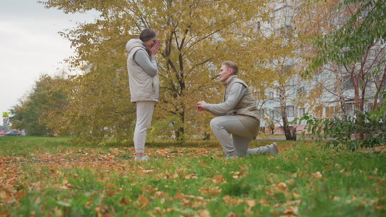 Young adult prepares to kneel and propose to fiancee in outdoor park as he reacts with surprise, surrounded by autumn leaves and trees with car and signpost visible in distant background