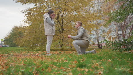 young adult prepares to kneel and propose to fiancee in outdoor park as he reacts with surprise, surrounded by autumn leaves and trees with car and signpost visible in distant background