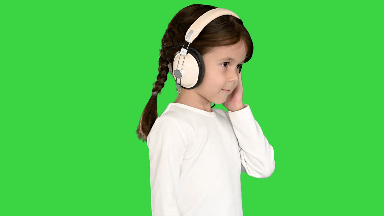 하 옷을 입은 귀여운 소녀가 한 손으로 헤드폰을 들고 녹색 스크린, 크로마 키를 는 동안 음악에 머리를 흔들고 있습니다.