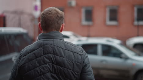 over shoulder shot of man wearing black winter coat and blue jeans walking toward parked car on urban pavement with red brick building in background during cold city day scene with subtle motion