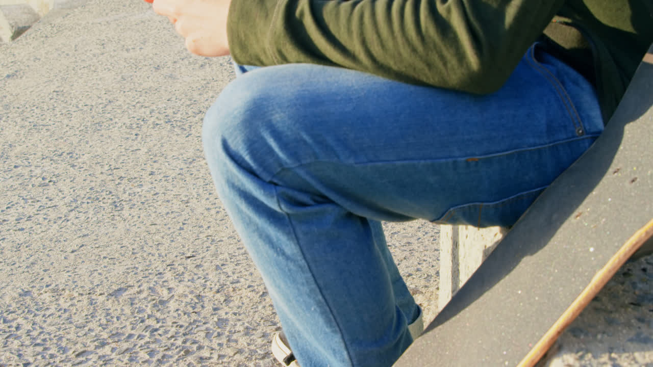 vista lateral de un joven caucásico enviando mensajes de texto en el teléfono móvil mientras está sentado en el pavimento 4k