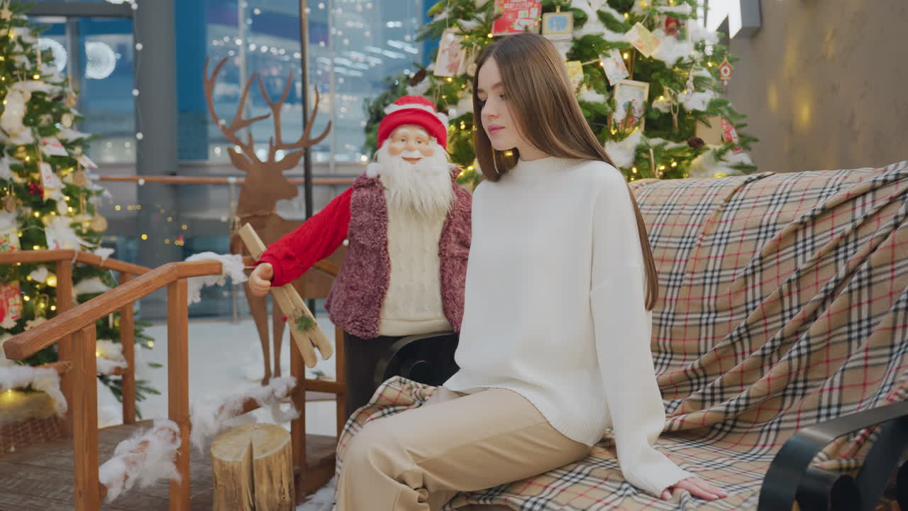 Young woman in brown trousers and white top sits on chair decorated with Christmas decor, enjoying festive ambiance with Christmas trees and lights in background