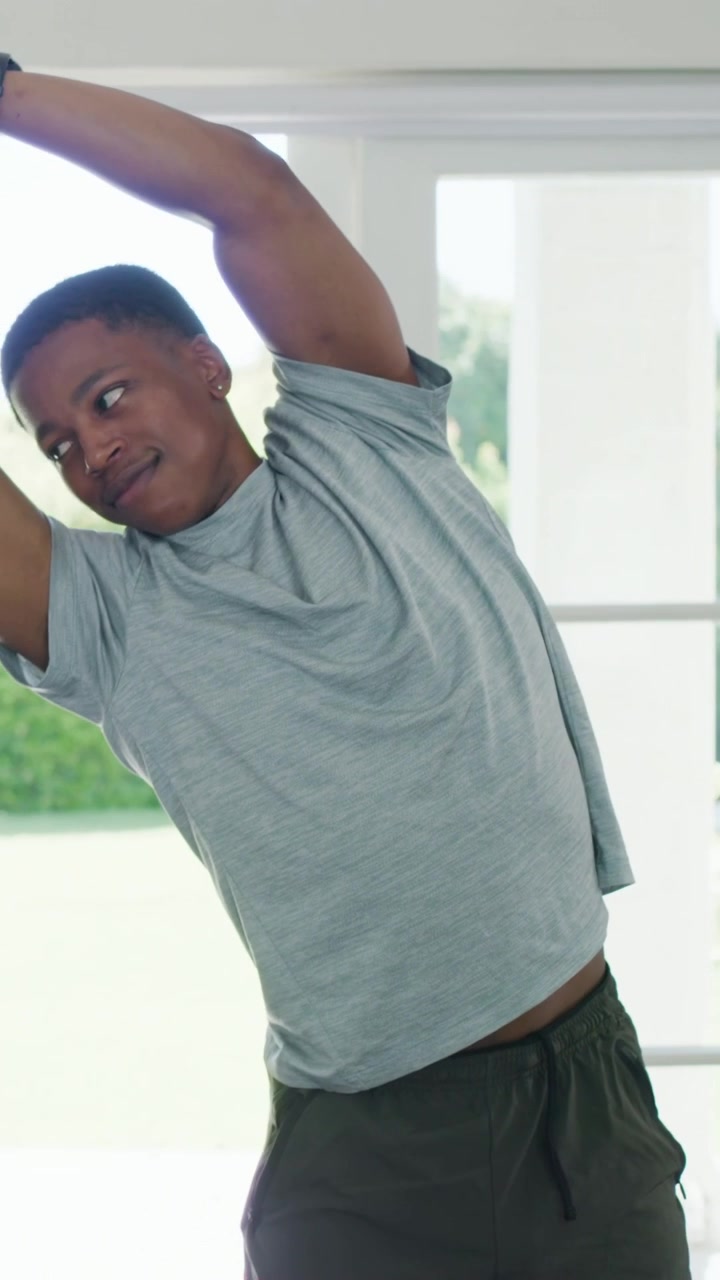 Vertical video: Stretching man in grey tee clasping hands overhead, tilting by glass doors, watch