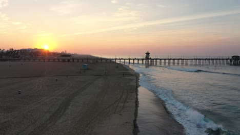 A-stunning-4k-view-of-the-pier-at-sunrise,-waves-crashing-,-and-surfers-surfing-in-Surf-City-California-USA