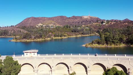 Aerial-People-Walking-Over-The-Dam-At-The-Hollywood-Reservoir-In-The-Hollywood-Hills-With-Hollywood-Sign-Distant