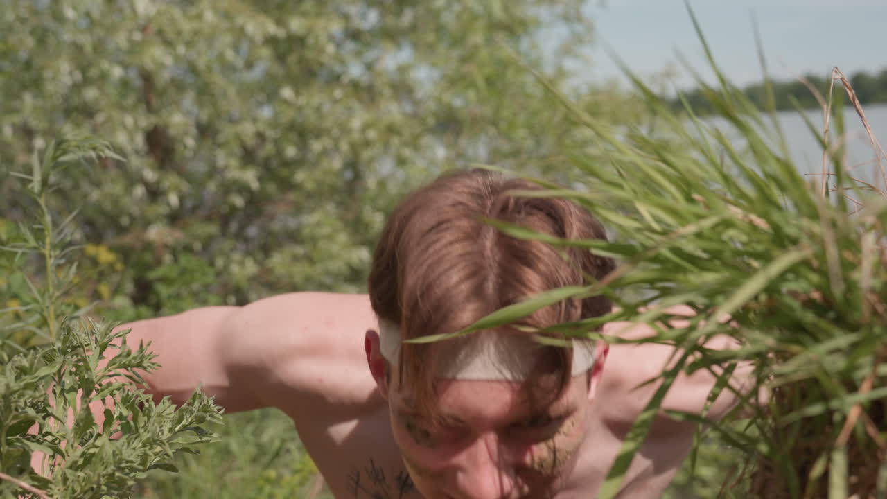 shirtless tourist with camouflage face paint and headband positions hands on grassy riverbank ready to perform push up exercise under bright daylight surrounded by lush greenery and flowing water