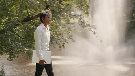 Tracking-shot-of-man-walking-at-park-carrying-a-laptop-for-online-remote-work