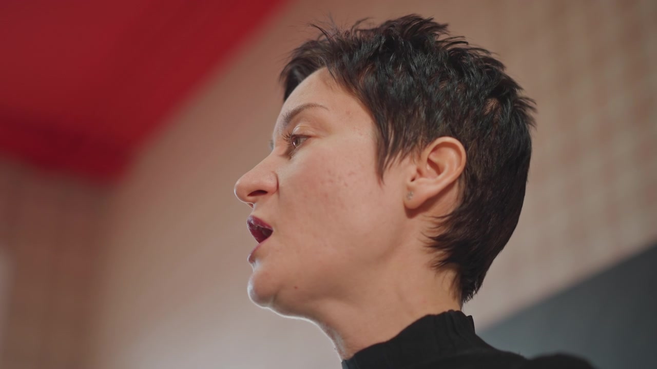 Vocal coach in una calda sala prove, primo piano di profilo di una donna con capelli corti che pratica il fraseggio sotto un soffitto rosso, concentrandosi su risonanza, dinamiche ed espressione emotiva, atmosfera di prova espressiva