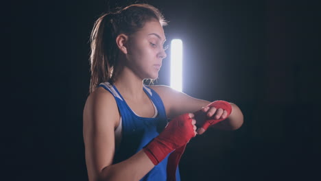 KSrendny-plan-beautiful-athletic-bride-boxer-in-blue-clothes-reels-red-bandages-on-the-garment-of-the-hands-of-a-female-fighter-on-a-dark-background.-steadicam-shot