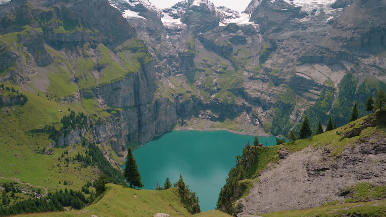 Lago alpino turchese circondato da ripide montagne rocciose coperte di neve e prati verdi, ripresa con movimento verticale verso l'alto.