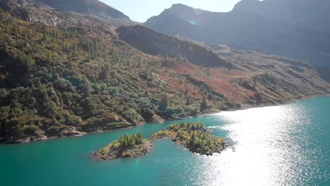 Aerial-flyover-over-sunlit-forested-islands-in-the-turquoise-waters-of-Lac-de-Salanfe-in-Valais,-Switzerland-on-a-sunny-autumn-day-in-the-Swiss-Alps-with-a-pan-down-view-from-the-mountains