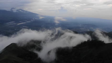 Mysterious-aerial-shot-flying-over-mountains-covered-with-fog,-white-clouds-at-sunrise,-a-sleeping-city-in-the-valley-at-dawn