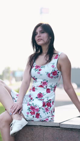 wonderful round motion long haired brunette girl in summer dress and sneakers sits on grey concrete parapet