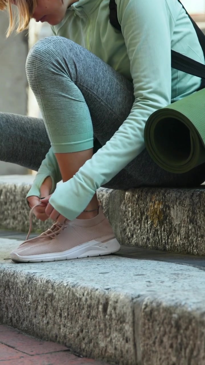Video verticale: Donna con felpa color menta che appoggia il piede su un gradino e allaccia scarpe rosa, con uno zaino e un tappetino da yoga.
