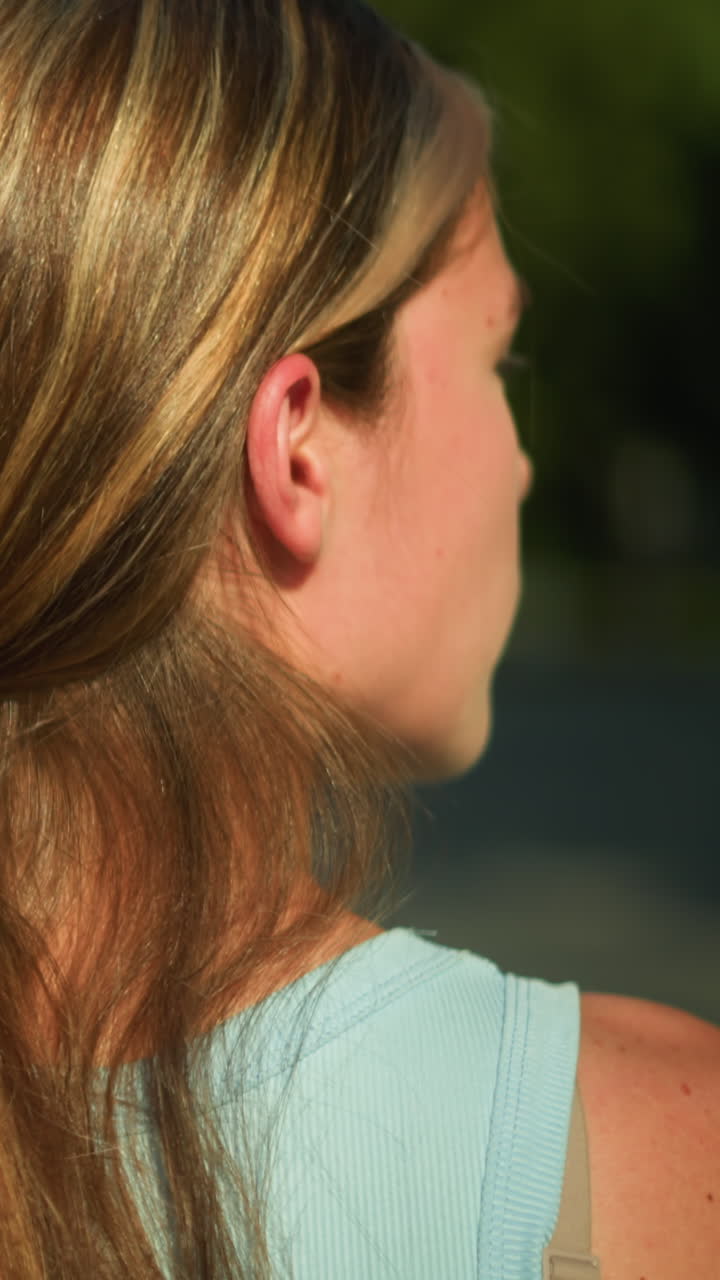 vista de atrás de cerca de una joven con el cabello largo atado hacia atrás usando un clip negro, mirando hacia la izquierda, con un fondo al aire libre borroso con vegetación suave y luz solar