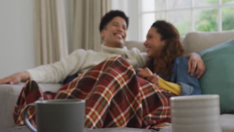 video of happy biracial couple siting on sofa under blanket and drinking coffee