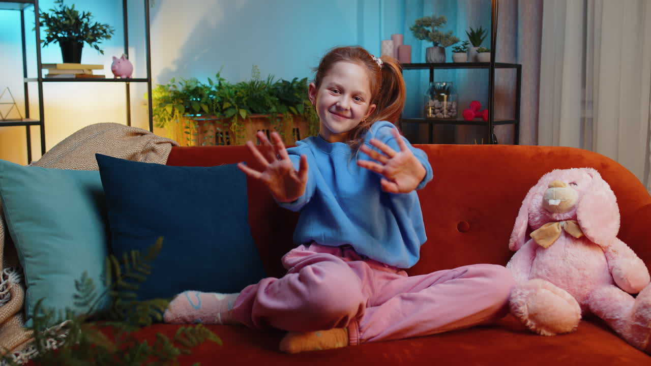 Child kid girl smiling friendly at camera waving hands gesturing hello hi greeting at home on couch