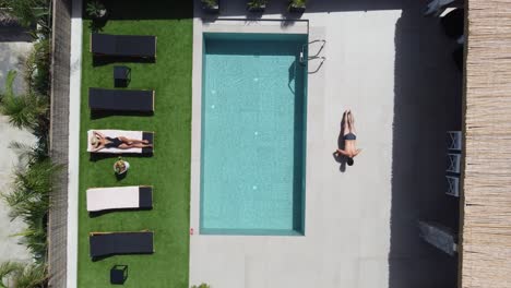 drone flying over an outdoor pool in bird's eye view - woman in swimming costume lying on sunbed man doing gymnastic exercises near pool edge - villa in greece crete with palm trees