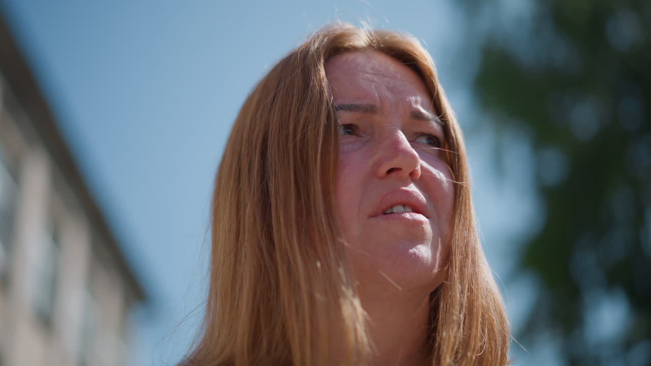 Close up of sad woman looking unsettled outdoors under bright sky with blurred green trees, emotional expression reflecting loneliness, contemplation, and life reflection in soft natural daylight